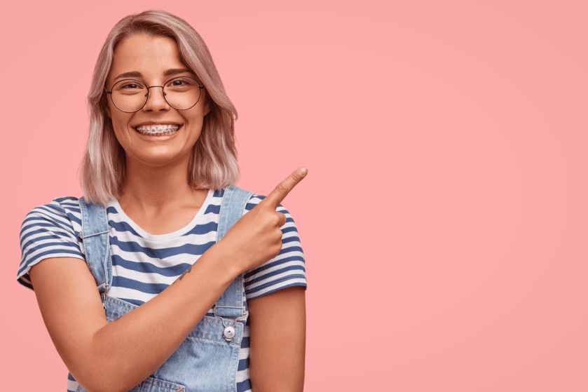 Teenage Girl on a pink background with braces, pointing at the important information of seeing an orthodontist.