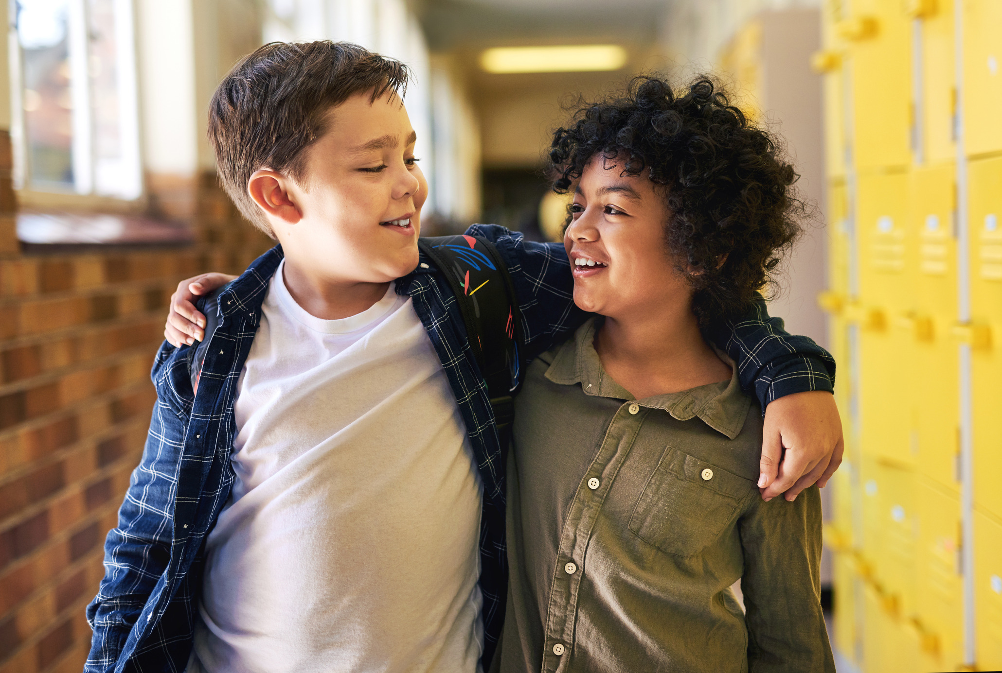 Two middle school aged boys, walking down the school hallway together with their arms around each other.