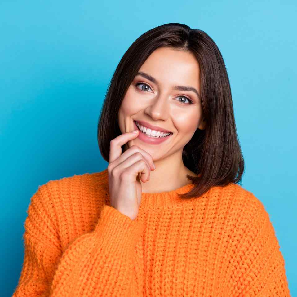 A young woman smiling. She just finished orthodontic treatment and has no appliances on. She is wearing an orange sweater and is on a blue background.